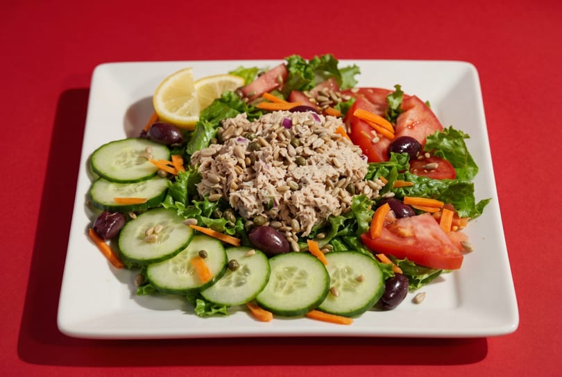 Tuna salad with lettuce, tomatoes, cucumbers, olives, sunflower seeds, and a lemon wedge.