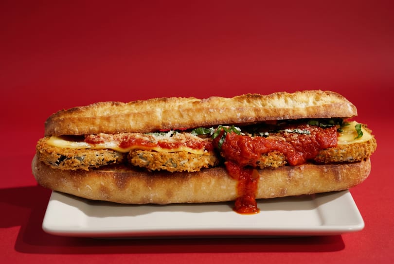 A large fried eggplant parmesan sandwich with marinara sauce and cheese on a white plate.