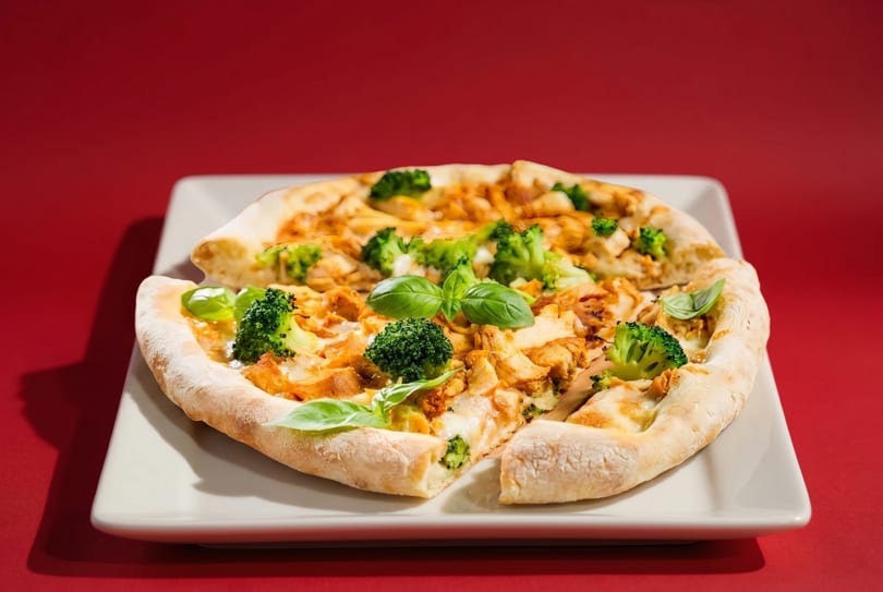 Chicken and broccoli pizza with basil on a white plate against a red background.