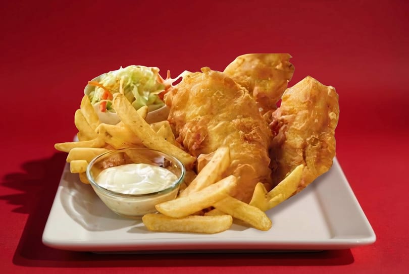Golden-brown fish and chips with coleslaw and tartar sauce on a white plate against a red background.